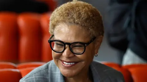 Getty Images This is a head and shoulders pic of Karen Bass who is smiling broadly and wearing a grey, checked suit. The background, slightly out of focus, appears to be red seating