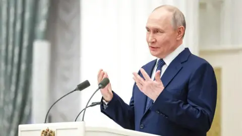 Reuters President Putin wearing a white shirt, dotted blue tie and blue suit, gestures with his hands as he speaks at a white podium with two microphones. A light green patterned curtain stands to the left of the picture.