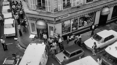 JACQUES DEMARTHON/AFP via Getty Images A black and white photo showin a restaurant surrounded by emergency vehicles