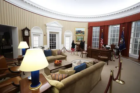 Talia Sprague/Bloomberg via Getty Images Stuffed chairs, lamps and dark wood furniture fill a replica of the Oval Office, with parts of the space sectioned off by red velvet ropes. Two people example the space.