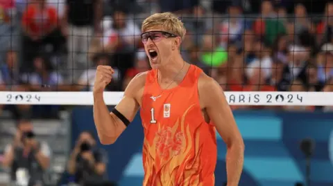 Reuters A male volleyball player wearing an orange singlet and tinted glasses, pumping his right fist and cheering, while standing in front of a volleyball net with a blurred crown in background.