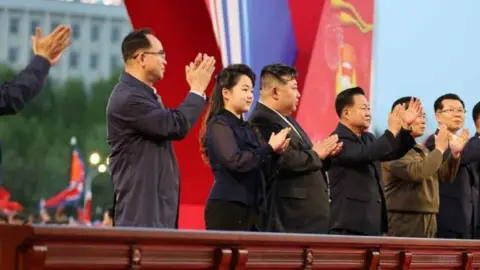 News1 With her long hair tied back, in a black see-through blouse, revealing her arms, Kim Ju Ae stands next to Kim Jong Un, in a black suit and white shirt, and several officials applauding the completion ceremony of Pyongyang's Vanguard Street development, with flags, lights, trees and a building in the background.
