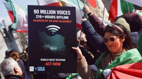 SOPA Images/LightRocket via Getty Images A woman a at a street protest, wrapped in an Iranian flag, holds up a placard. It reads: 90 million voices, 216 + hours offline, silenced by IRGC blackout. Other people holding Iranian flags can be seen in the background. 