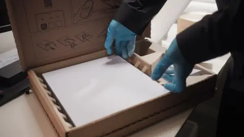 BBC A white rectangular Starlink device rests inside a brown cardboard box. The hands of a person wearing thin blue plastic gloves and dark clothing are touching it at the edges, as if lifting it out of the box.