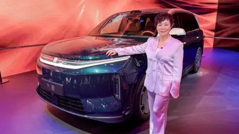 Stella Li is wearing a silky lilac suit and leaning against a BYD SUV while speaking to the BBC at the Beijing auto show.
