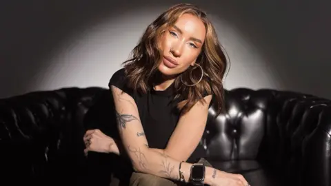 BBC Katya has long mid-brown hair and blue eyes. She is wearing big hoop earrings and has her nose pierced. She has tattooes in the shape of plants and butterflies down one arm. 