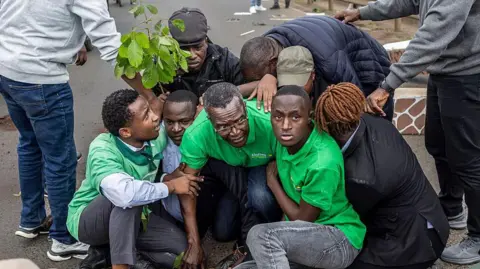 AFP via Getty Images Eight people crouched in a huddle and they are being detained. There is a person standing on either side, keeping them down