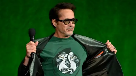 Getty Images Robert Downey Jr on stage at CinemaCon, opening his jacket to reveal a T-shirt showing the masked face of his character Doctor Doom