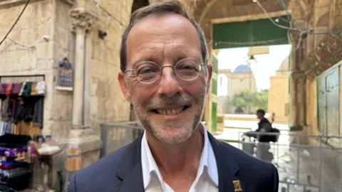 Maarten Lernout/BBC Man with small round metal framed glasses and a grey suit with a weight shirt and a little gold badge on his lapel. He is smiling in the background there is a barrier 