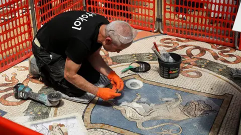 Mondadori Portfolio via Getty Images A restorer kneels in front of a mosaic of a bull. He is wearing orange cloves and a black T-shirt. An orange partition is set up around him to separate him from the public.