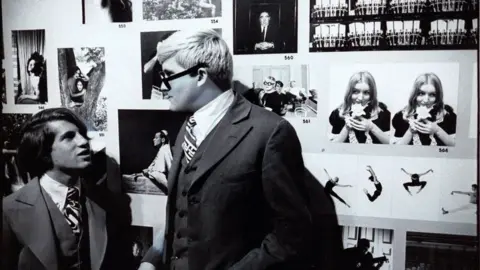 Getty Images David Hockney and Peter Schlesinger. This is a black and white photo of Hockney and his former boyfriend. They are in a gallery surrounded by black and white photos. They are captured while having a conversation.