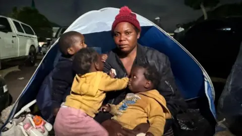Thuthuka Zondi / BBC Esnat Joseph, wearing a maroon woollen bobble hat and black jacket, sitting in front of a tent with her baby triplets - two dressed in yellow hoodies and one black.