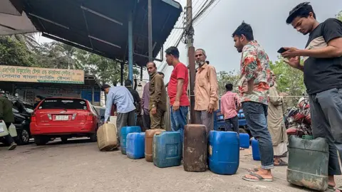 Getty Images Motorists wait in line to purchase fuel at a petrol station in the Mohammadpur area of Dhaka, Bangladesh. Several people are standing in a queue with various petrol cans waiting to be filled