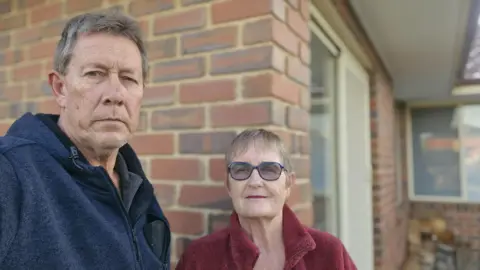 Supplied A middle aged man and woman wearing glasses stare at the camera in front of a brick house
