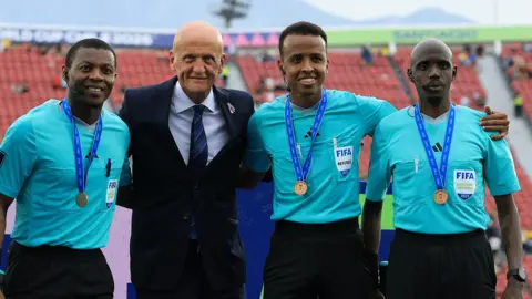 Getty Images Pierluigi Collina, wearing a dark suit, blue striped tie and white shirt, is flanked by Omar Artan and two other officials who are all wearing light blue shirts and dark shorts. All three officials have medals around their necks on blue ribbons, and a sparsely populated stand is seen out of focus in the background