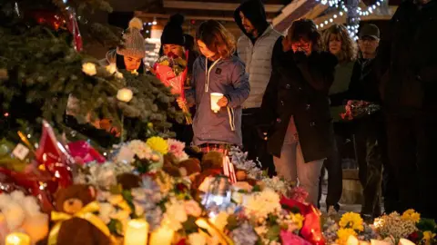 AFP via Getty Images Community members mourn during a candlelight vigil for the victims of Tumbler Ridge Secondary School where a mass shooting took place a day earlier in the small town of Tumbler Ridge, British Columbia, on February 11, 2026. A pile of flowers and candles can be seen at the forefront of the image, while several people look down in the background towards the vigil with sombre expressions. 