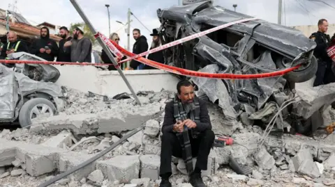 EPA A man sits on the rubble in front of a mangled car