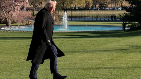 REUTERS/Kylie Cooper US President Donald Trump walks across the South Lawn with a black gate and a fountain in the background. He is wearing black gloves, a long black coat and his looking down. His hair is blowing.
