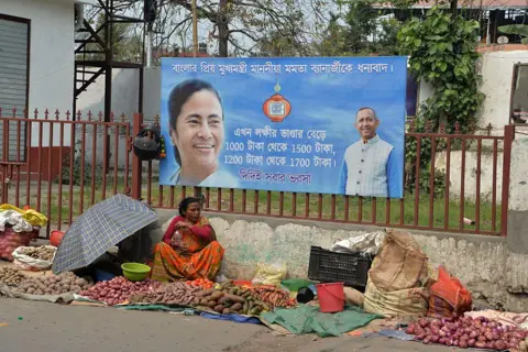 NurPhoto via Getty Images Women vegetable sellers wait for customers at a market area on their roadside stalls near a poster of West Bengal Chief Minister Mamata Banerjee's recent Laxmi Bhandar (monthly allowances to West Bengal women) in Siliguri, India, on 