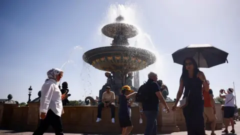 Reuters People walk around a fountain, with blue sky and bright sunshine in the background. One woman is holding an umbrella for shade; other people are wearing sunhats and carrying bottles of water.