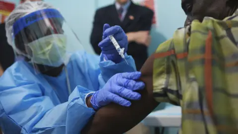 Getty Images A nurse in blue scrubs, visor and face mask administers an injection on the first day of the Ebola vaccine study being conducted at Redemption Hospital, Monrovia, Liberia - 2015.