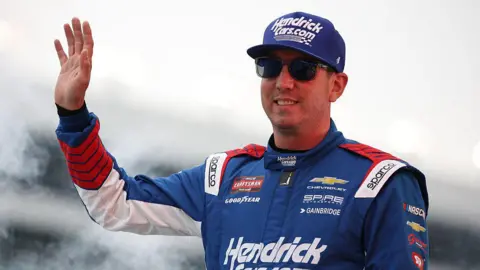 Getty Images Kyle Busch, in a blue, red and white Nascar driver's jumpsuit and matching blue cap, as well as sunglasses, smiles and waves. 