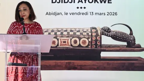 AFP via Getty Images Ivorian Minister of Culture and Francophonie Françoise Remarck talks at a podium in front of a picture of the ornately carved wooden drum