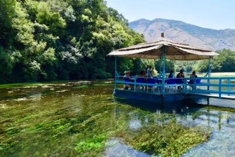 Amber Robertson A veranda on a river in Albania. The water is crystal clear and you can see the green plants on the river floor. It is hot and sunny.