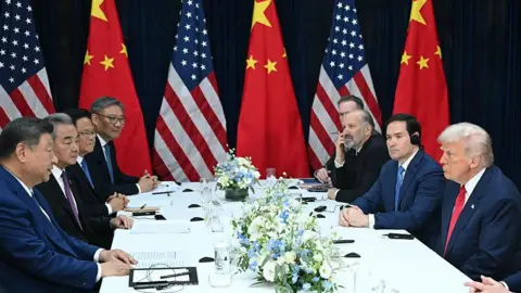 AFP via Getty Images President Xi facing Trump across a meeting table, flanked by their countries' representatives for bilateral talks. Flags of the US and China are displayed in the background.