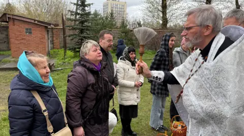 BBC Priest dressed in a white robe giving his blessing to churchgoers.