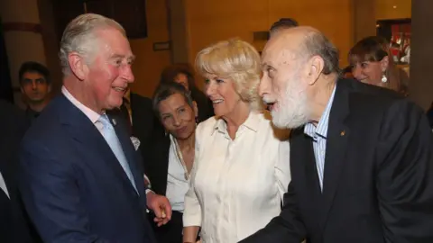 Getty Images The King on the left of the image, wearing a navy blue suit, pink shirt and pale blue tie shakes hands with Petrini, who is on the right in a dark blue suit and blue and white striped shirt. The Queen is in the middle smiling in a white dress.