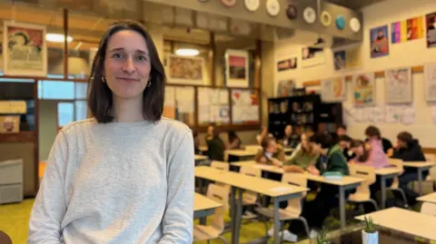 BBC A brunette woman with a bob smiles at the camera in front of a classroom of teenagers. She is smiling and wearing a grey jumper. There are about a dozen children in the class