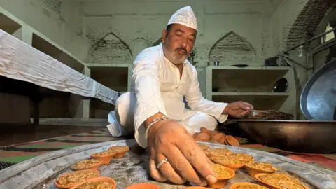 Aman/BBC Murtuza Hussain Raju, wearing a white kurta and a skull cap, prepares kebabs while sitting on the floor. 