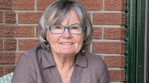 Supplied/Elizabeth Doran An image of Elizabeth Doran. She is smiling and has a short grey bob with bangs, blue-framed eyeglasses and dangling earrings. She is wearing a collared linen grey-mauve shirt, and behind her is a brown brick backdrop. 