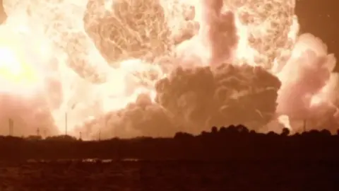 SPACEFLIGHTNOW A vast orange fireball erupts from the horizon at Cape Canaveral, photographed from across the water at night. The blast fills the upper two-thirds of the frame in churning, billowing clouds of incandescent gas — bright white at the heart of the fireball, fading through gold and amber to deep orange at the edges. A second, equally violent plume mushrooms upward at the centre, lifting smoke and debris into the sky.