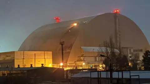 IAEA HANDOUT/EPA-EFE/REX/Shutterstock The large curved metal shield over reactor four, pictured in low light, with flame emerging from a hole in one side where it was hit by a drone on 14 February 2025. There are red lights on top of the dome, and there is yellow lighting around the lower buildings close to it.