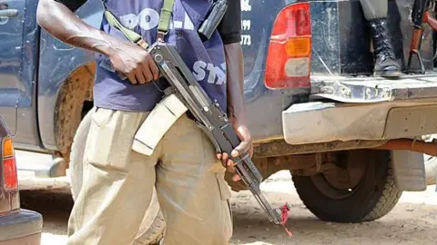 AFP via Getty Images Armed police officer stands beside the back of patrol trucks, holding a rifle in Jos, Plateau State.