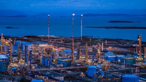 Getty Images A night-time view of a gas processing plant in Tysavaer, Norway. It shows a wide view of the plant with various buildings and processing barrels. Three tall columns can be seen from the distance emitting flares. The plant overlooks the sea.