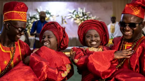 AFP via Getty Images Twin brothers Kehinde and Taiwo Oguntoye carry Kehinde and Taiwo Adediran. Both couples wear red traditional formalwear.