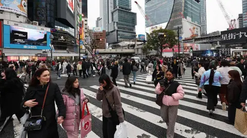 NurPhoto via Getty Images A crowded Shibuya crossing in the day, with tall buildings seen behind