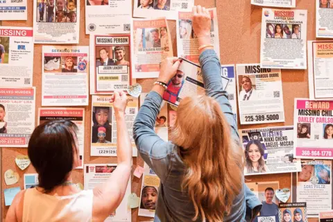 CrimeCon and RenownedPhotos Two women look at a board filled with missing people posters