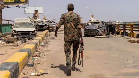 Avaaz via Getty Images A lone soldier walks across a bridge. Bombed-out cars and rubble can be seen on the bridge.