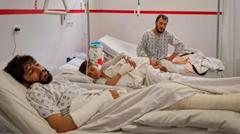 Reuters Two dark-haired men and a boy lie and sit in their hospital beds. They are wearing hospital gowns. One man has a large dressing on his arm, and the boy has a dressing on his hand.