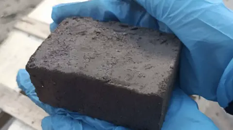 The Earth Prize A close-up shot of the large, dark brown brick held by a pair of hands in blue surgical gloves.