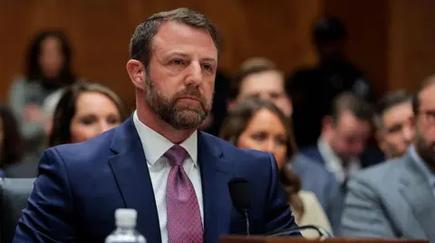 Getty Images Mullin sits in a blue suit and red patterned tie and looks ahead while speaking from a microphone