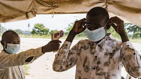 AFP via Getty Images man on left wearing a mask and holding up a contactless thermometer to a man who is putting on a mask. There are standing in a tent
