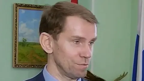 SEIMTVKursk Igor Khudokormov is photographed against a green wall with a painting showing fields and trees in the background. He is wearing a navy suit jacket and light blue shirt.