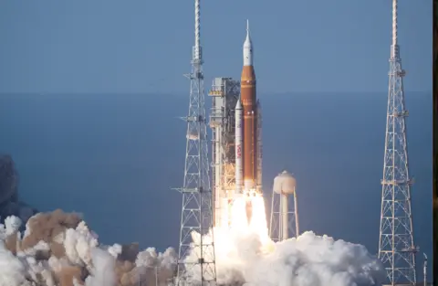 NASA A tall white and orange rocket is blasting off from a coastal launch pad. Flames and bright white exhaust pour from its base, creating a wide, glowing tail that hides the ground in thick clouds of smoke. The rocket is pencil‑thin and points straight up into a clear blue sky, with the sea faintly visible behind it. Slender white booster rockets cling to either side of the central orange core. To the left and right stand two latticework metal towers, like giant scaffolding poles, framing the rocket. A rounded white tank sits nearby on spindly legs, partly lost in the steam. The overall impression is of immense power and light as the vehicle climbs away, leaving a boiling, churning cloud where it stood.