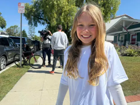 A 10-year-old girl with blond hair and white t-shirt smiles for the camera while her dad is interviewed on camera in the background