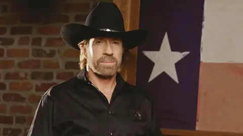 Getty Images Chuck Norris wears a black shirt and a cowboy hat while posing for a photo. Behind him is a Texas flag and a brick wall